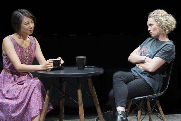 Two women sit opposite each other at a small round table with two black mugs. One woman wears a pink dress and holds a mug, while the other, in a t-shirt and jeans, sits with arms crossed, appearing upset.
