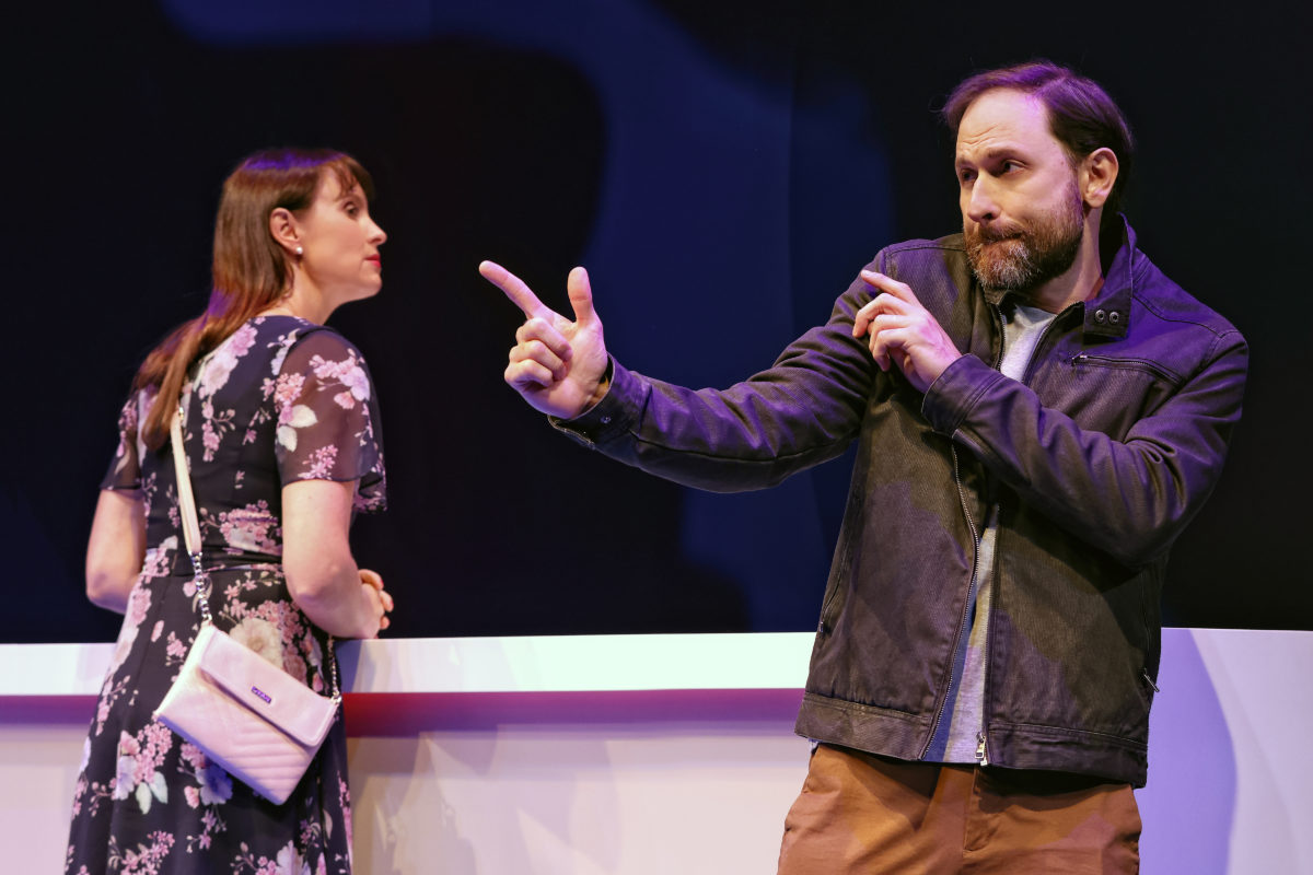 A woman in a floral dress looks away whilst a bearded man in a jacket points his fingers like a pretend gun, striking a playful pose on stage with a dark background behind them.