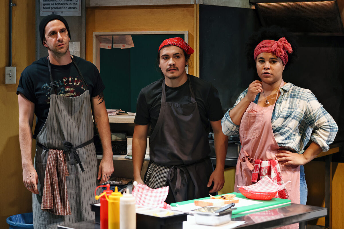 Three people in aprons and headbands stand behind a kitchen counter with food baskets and condiments, looking towards the camera with serious expressions. The kitchen setting includes shelves and cooking equipment in the background.