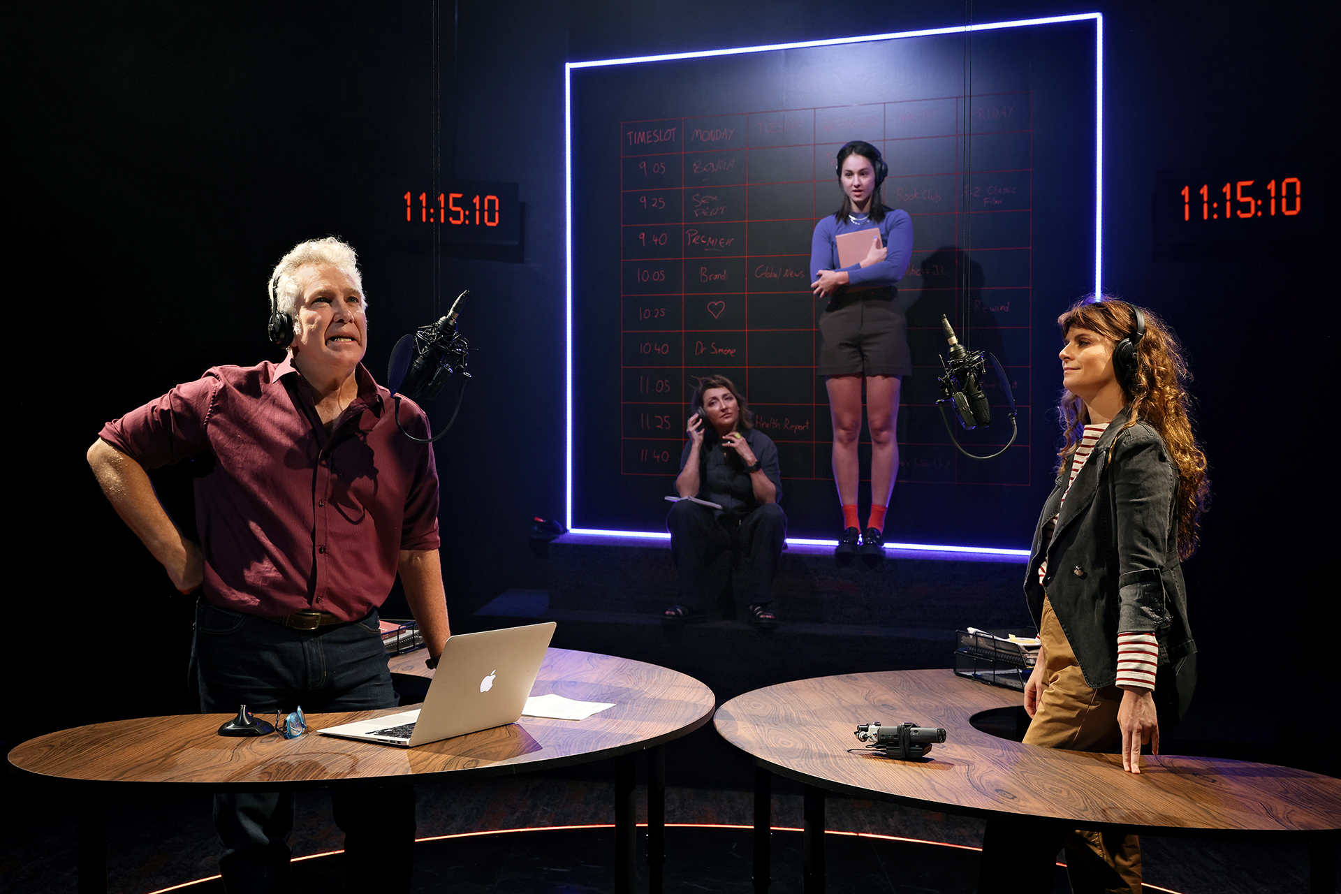 Three people stand at round desks with microphones, while a fourth person stands in front of a large board with notes and a neon outline. Two digital clocks show the time as 11:15:10 in a dark, studio-like setting.