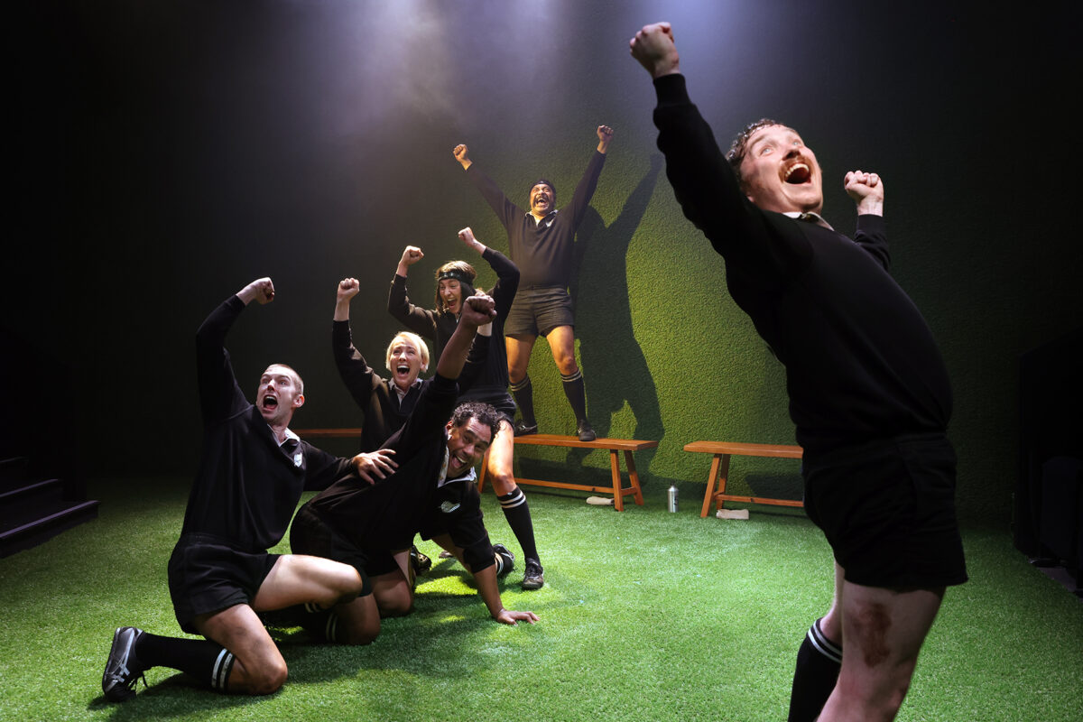 Six people wearing black rugby kits celebrate energetically on a green pitch, some kneeling and others standing with fists raised in victory, under dramatic stage lighting. Benches are visible in the background.