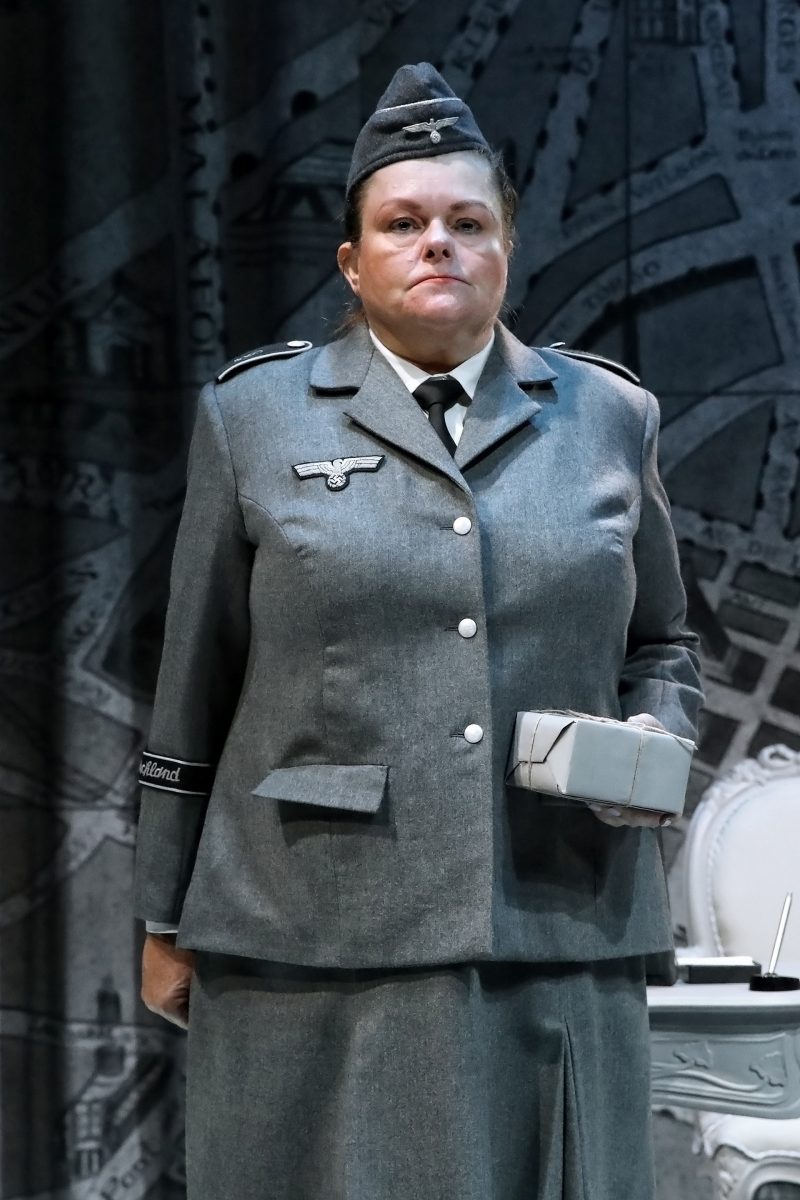 A person in a grey military-style uniform with insignia stands in front of a map-themed backdrop, holding a small, wrapped box.