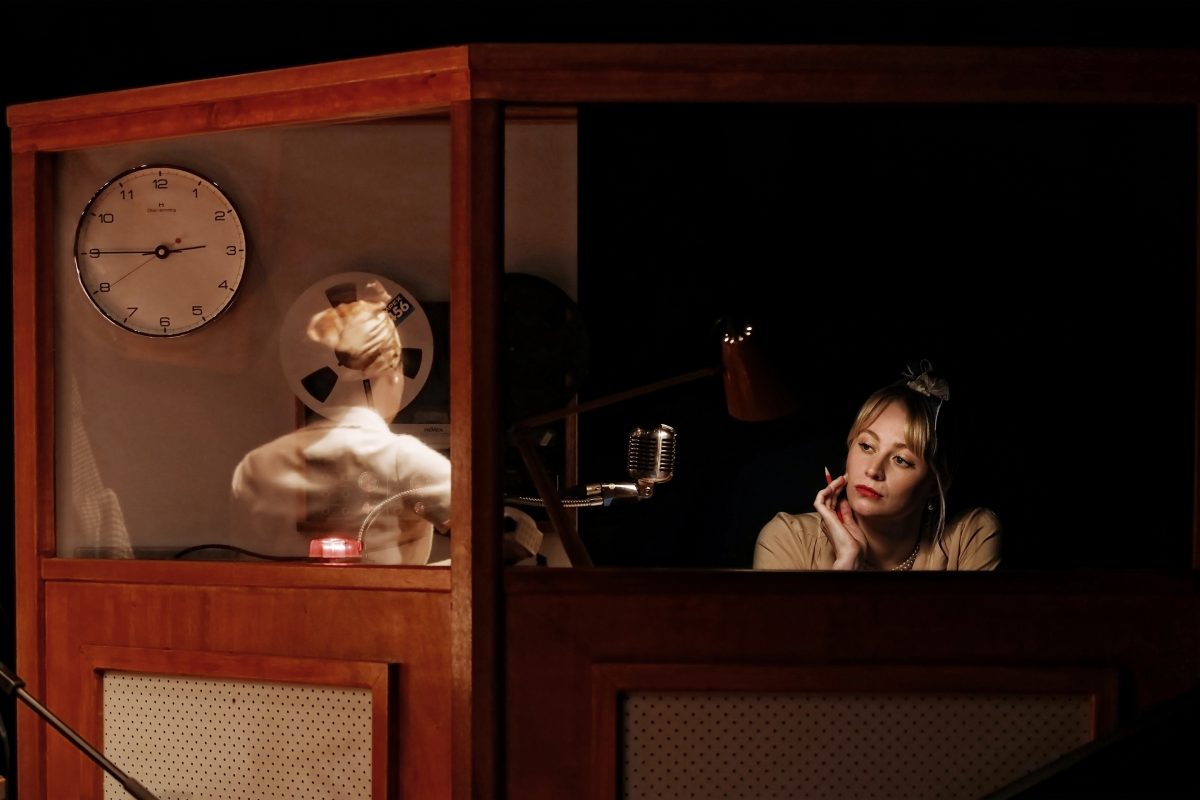 A woman sits thoughtfully in a dimly lit recording studio booth, resting her chin on her hand near a vintage microphone. A clock and reel-to-reel tape recorder are visible behind a glass partition.