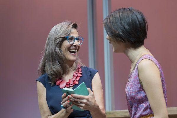 Two women smiling and talking together. One is holding a mobile and wearing glasses and a bold red necklace, while the other has short dark hair and is wearing a patterned sleeveless top. The background is softly lit in pink tones.