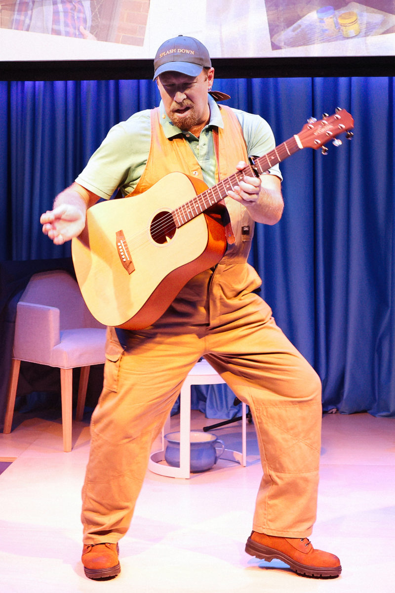 A man in orange overalls and a blue cap plays an acoustic guitar passionately onstage, standing in a dramatic pose with blue curtains and a white chair in the background.