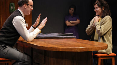 A man and a woman sit at a wooden table, facing each other; the man gestures with both hands, whilst the woman smiles with her hands clasped. Another woman stands in the background, watching them.