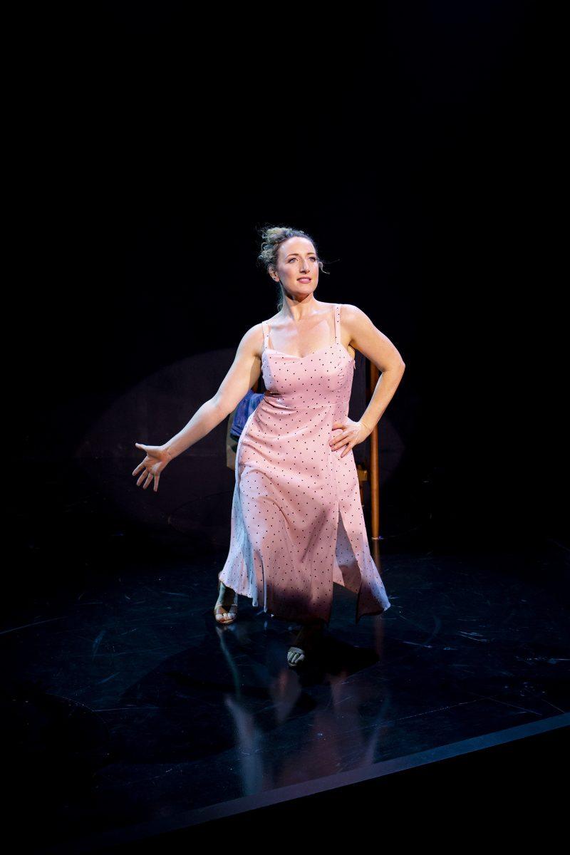 A woman in a light pink dress with small polka dots performs on stage under a spotlight, striking a theatrical pose with one hand extended and one knee bent, standing in front of a wooden chair.