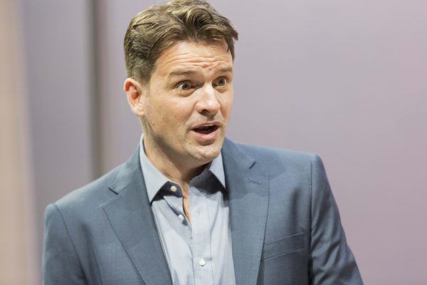 A man in a blue suit and light shirt is speaking or reacting, with a surprised or expressive facial expression, against a plain, blurred background.