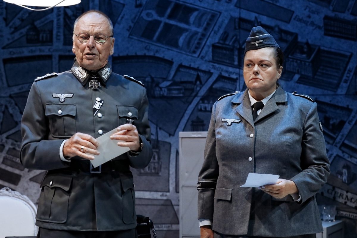 Two actors in Nazi military uniforms stand on stage, holding papers, against a blue-toned map backdrop. The man speaks whilst the woman looks serious and attentive. The scene appears to be from a theatrical performance.