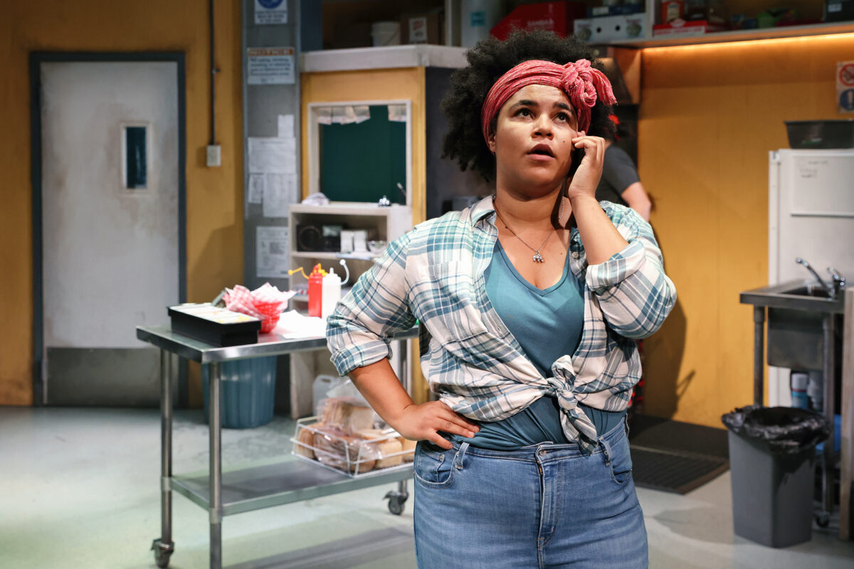 A woman with curly hair wearing a red headband, checked shirt, and jeans stands in a kitchen setting, holding a phone to her ear and looking concerned. The kitchen has shelves, stainless steel tables, and food containers.