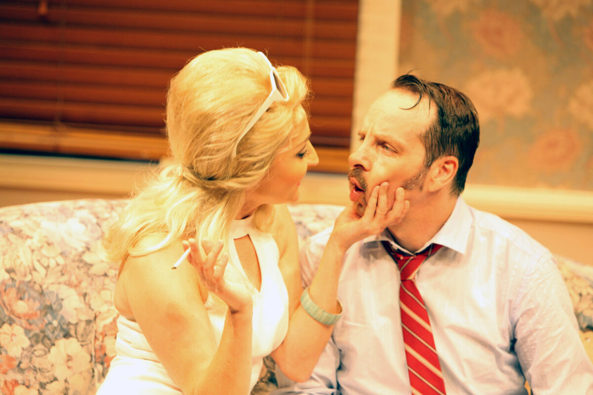 A woman with blonde hair and a white dress holds a cigarette and gently touches a man’s face as he looks at her, seated on a floral-patterned sofa in a warmly lit room with blinds and patterned wallpaper.