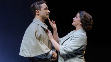 A woman smiles and gently touches a man's chin while holding his hands. Both wear vintage-style clothing and stand against a dark background, suggesting a scene from a play or performance.