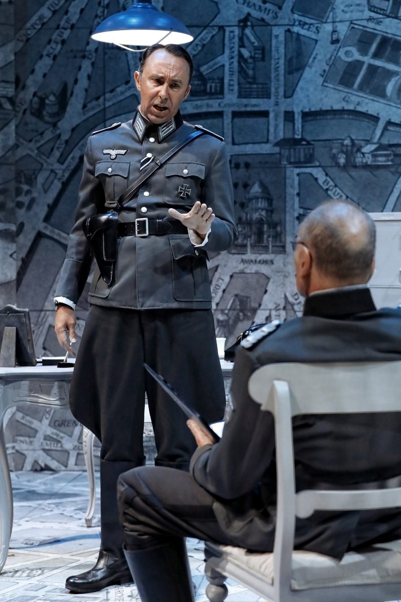 Two men in Nazi military uniforms are in a dramatic scene; one stands and gestures with his hand whilst speaking, and the other sits facing him, holding a folder. The setting includes a wall covered with maps and a desk.