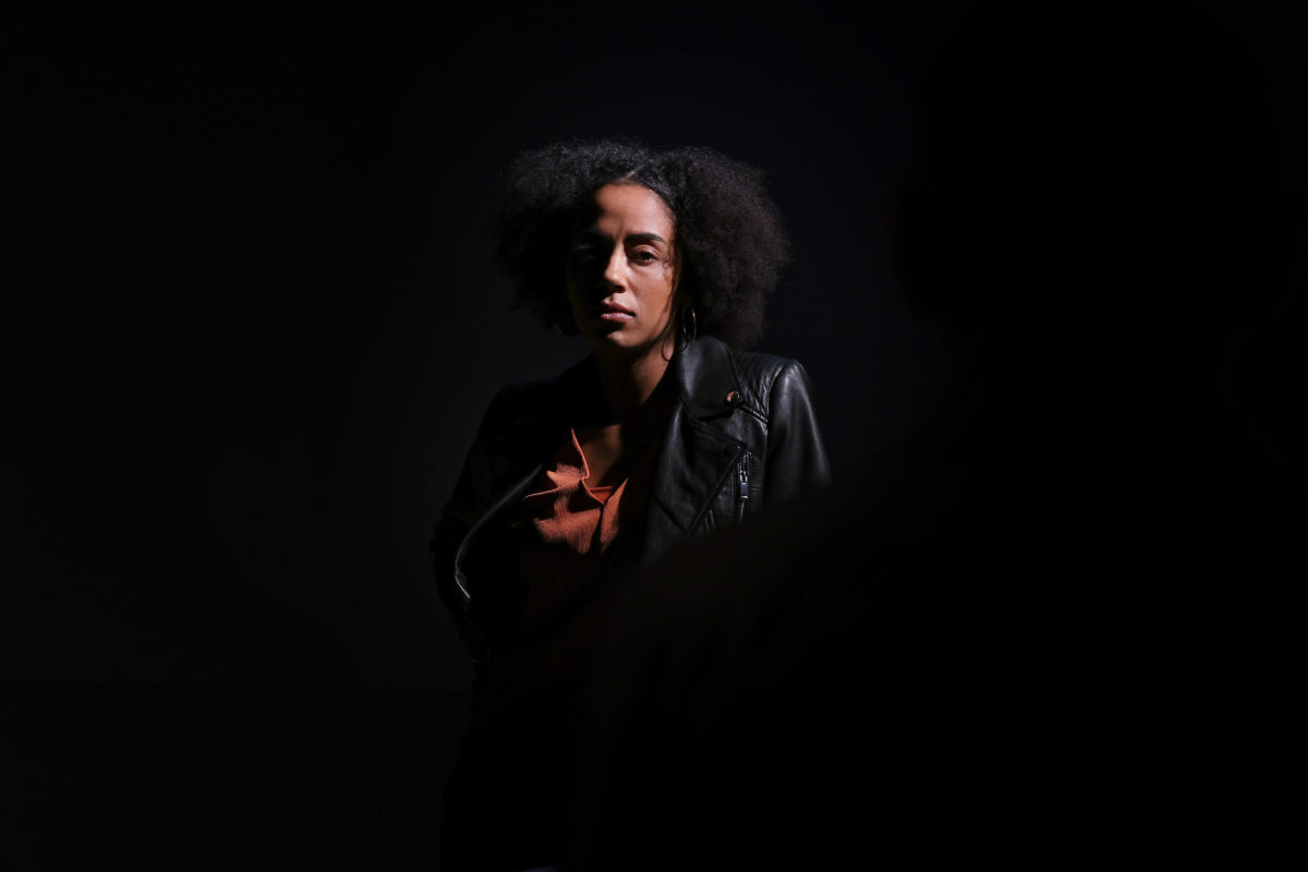 A woman with curly hair, wearing a leather jacket and rust-coloured shirt, stands in dramatic lighting with most of her face in shadow. A dark, blurry figure is visible in the foreground.
