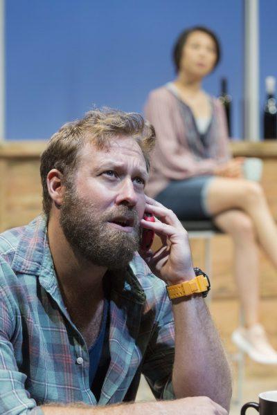 A bearded man in a checked shirt and orange watch talks on a red phone, looking concerned. In the background, a woman with short hair sits on a counter, slightly out of focus.