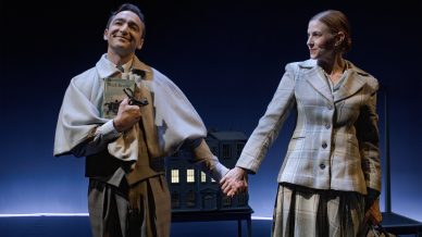 A smiling man holding a book and a woman holding a bag stand on stage, dressed in vintage-style clothing, holding hands in front of a model house against a dark blue background.