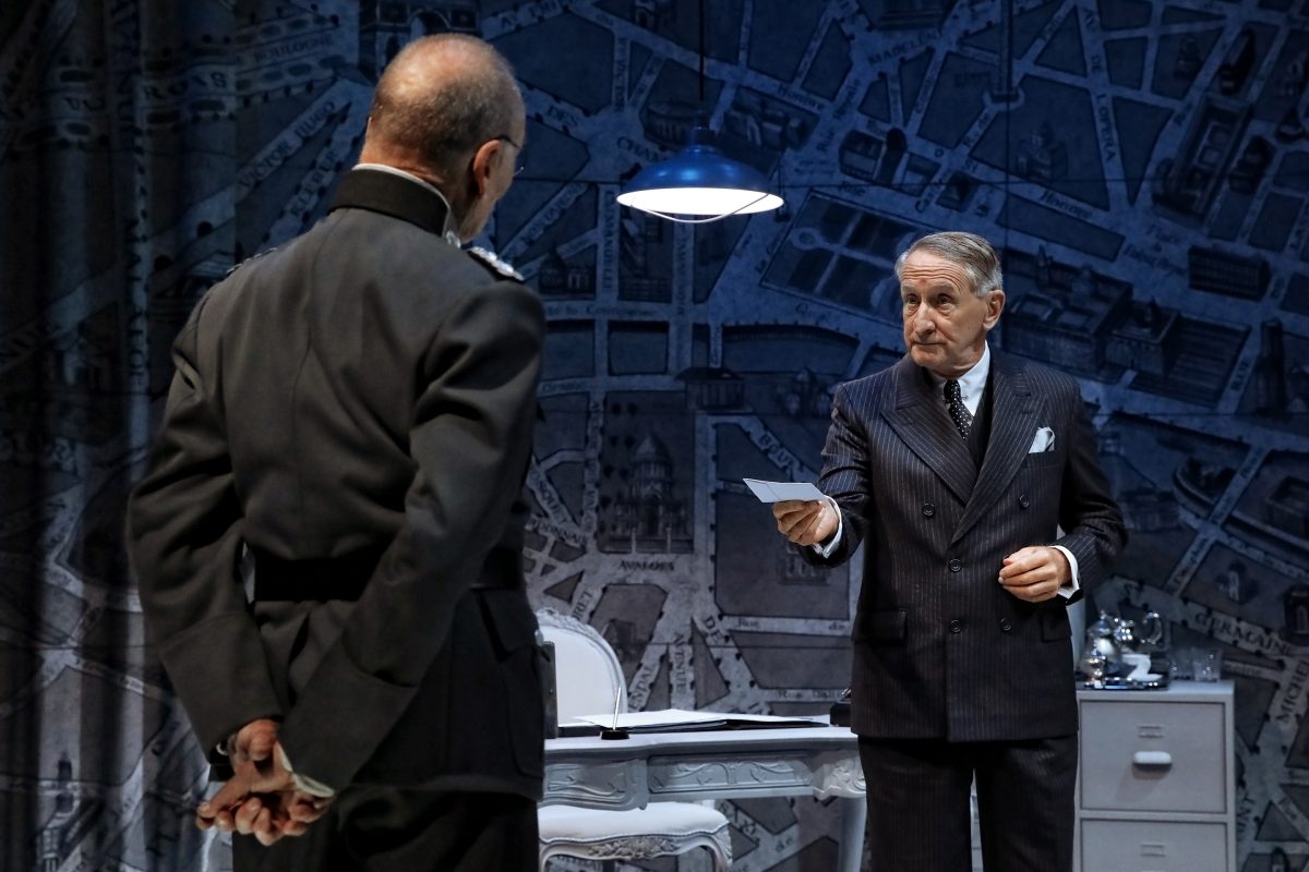 Two men in formal attire stand in a dimly lit office with a map wallpaper background; one faces away with hands behind his back, the other faces him, holding out a piece of paper.