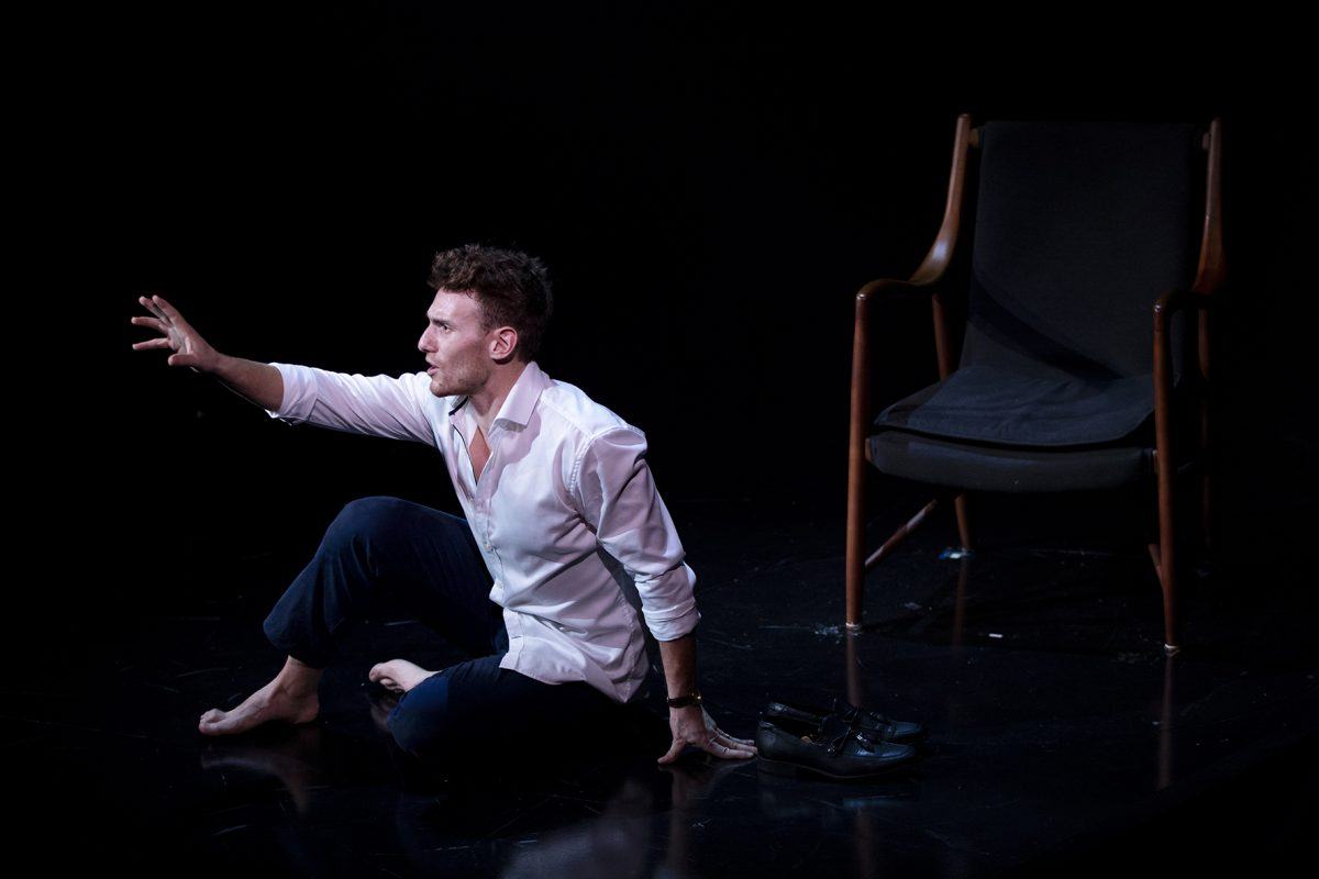 A barefoot man in a white shirt and dark trousers sits on the floor, reaching out with one hand. He faces forwards, with a pair of shoes beside him and an empty chair in the background, all against a dark backdrop.