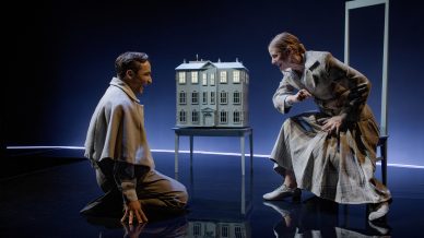 Two actors on stage face each other intensely; one kneels while the other sits and gestures towards a detailed dolls’ house model on a table between them. The scene is dramatic, with dim lighting and minimal set design.