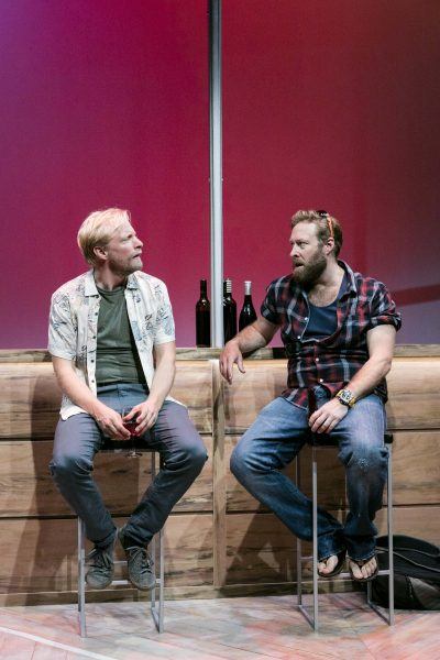 Two men sit on bar stools in front of a wooden bar with wine bottles on it. One man wears a patterned shirt and holds a glass, while the other wears a checked shirt, jeans, and flip-flops. The background is a gradient red.