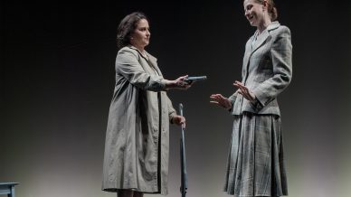 Two women stand on a reflective black stage. One, in a long coat, holds a rifle and offers a handgun to the other, who wears a checked suit and gestures with open hands, declining the weapon. Their reflections are visible on the floor.