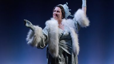 A woman in a glamorous blue vintage dress with white fur trim poses confidently onstage, wearing gloves and a feathered hat, with a bench beside her and a dark blue background.