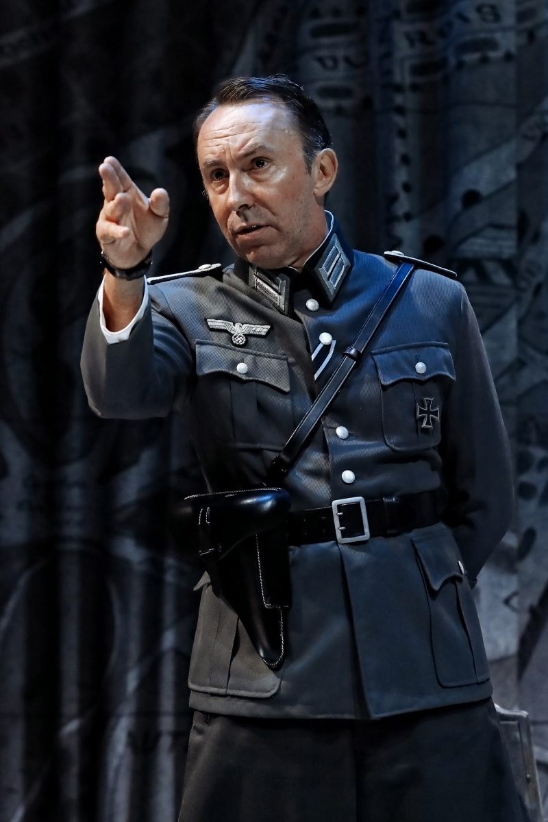 A man in a Second World War-era German military uniform stands with one hand pointed forward, as if giving a command. He has short hair and a serious expression, with a dark backdrop behind him.