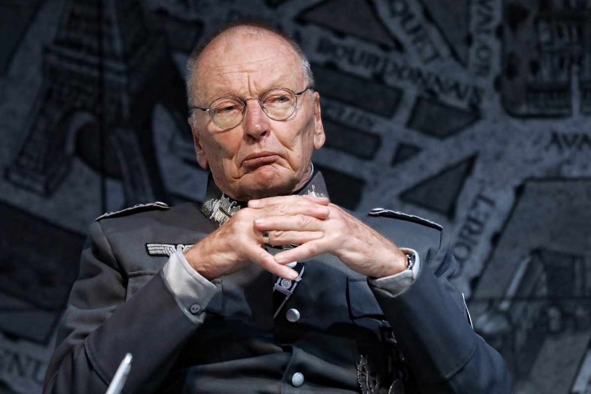 An older man in a grey military-style uniform with glasses sits with his hands clasped, looking stern. The background appears to be a map or diagram in dark tones.