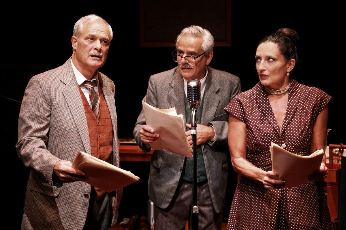 Three actors dressed in vintage clothing stand together, holding scripts and reading into an old-fashioned microphone, appearing to perform a radio play on stage. The setting is dimly lit with a dramatic atmosphere.