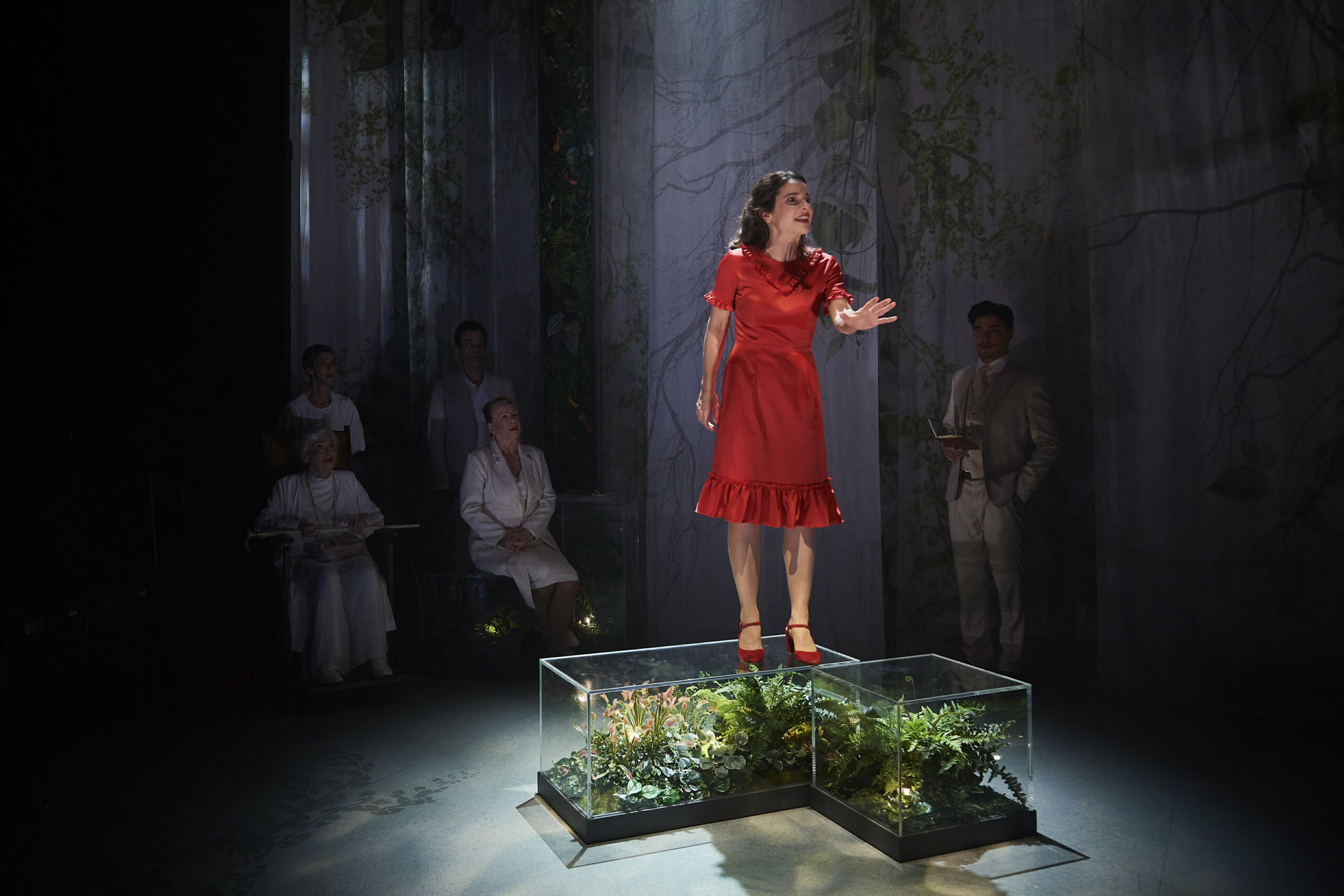 A woman in a red dress stands on a glass platform filled with plants, speaking or singing, while four people in white outfits watch her from the background on a dimly lit stage with a forest-like backdrop.