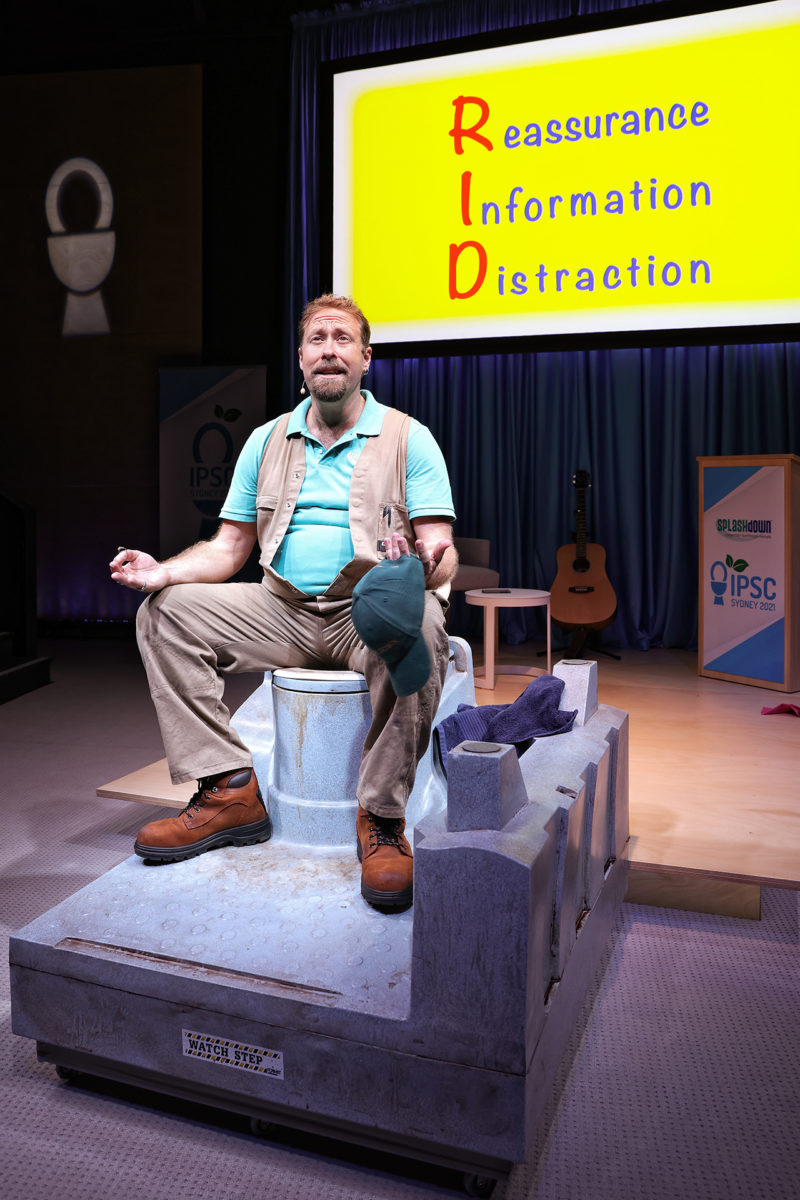 A man sits cross-legged on a stage prop resembling a giant latch, holding a green hat. Behind him, a screen displays RID: Reassurance, Information, Distraction in colourful text. A guitar and banners are in the background.