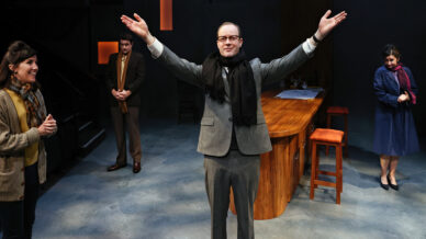A man in a suit and scarf stands centre stage with arms wide open, smiling. Three other people, two women and a man, stand around him, all appearing engaged. The setting is a modern bar with warm lighting.