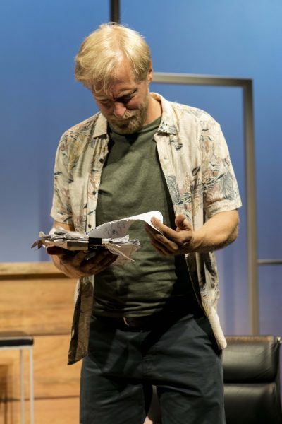 A man with blond hair and a beard, wearing a floral shirt over a green T-shirt, looks down sadly at a torn, crumpled stack of papers in his hands. The background includes a wooden table and a blue wall.