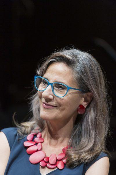 A woman with long grey hair, wearing blue glasses, red earrings, and a chunky red necklace, smiles while looking slightly to the side. She is dressed in a dark top with a blurred dark background behind her.