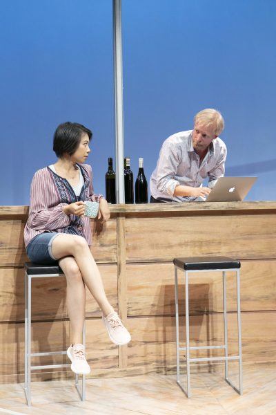 A woman sits on a barstool holding a mug, looking at a man using a laptop at a wooden bar. Three wine bottles are on the counter. The background is blue.