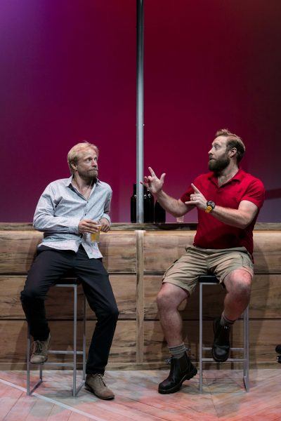 Two men sit on barstools in front of a wooden bar. One, wearing a light shirt and holding a drink, listens as the other, in a red polo and boots, gestures animatedly. The background is lit with a red gradient.