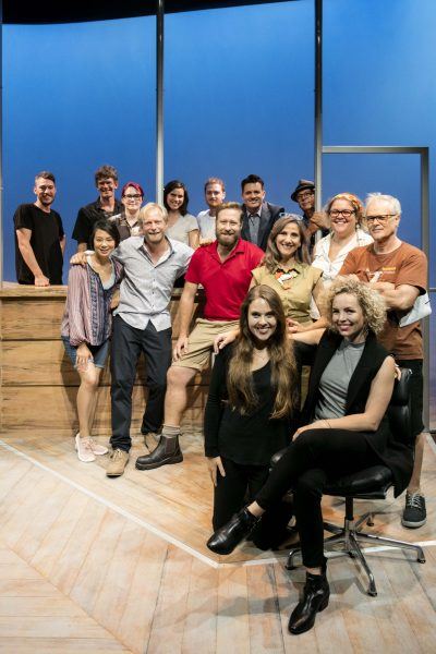 A group of 15 adults casually pose and smile together on a brightly lit stage set with blue walls and wooden floors. Some are standing, whilst others are seated or kneeling at the front.