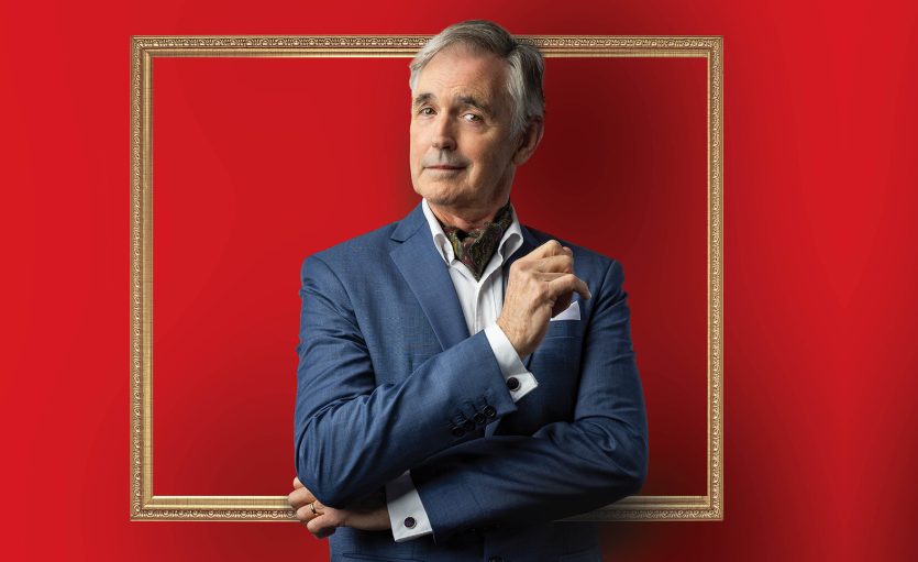 A confident older man in a blue suit poses with folded arms, standing in front of a red background with a gold picture frame around his upper body.