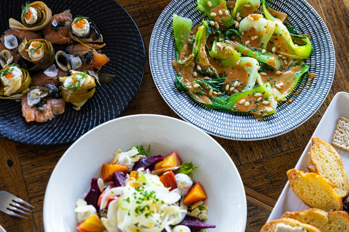Four plates of food on a wooden table: seafood starters, a green vegetable salad with a creamy dressing, a beetroot and fennel salad, and sliced toasted bread with biscuits.