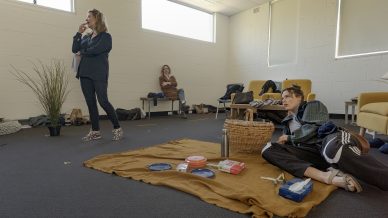 Three women are in a bright room with white walls. One stands, thinking, while two sit on the floor and yellow chairs. A brown blanket with picnic items and a basket is spread on the carpet. The atmosphere is casual and relaxed.