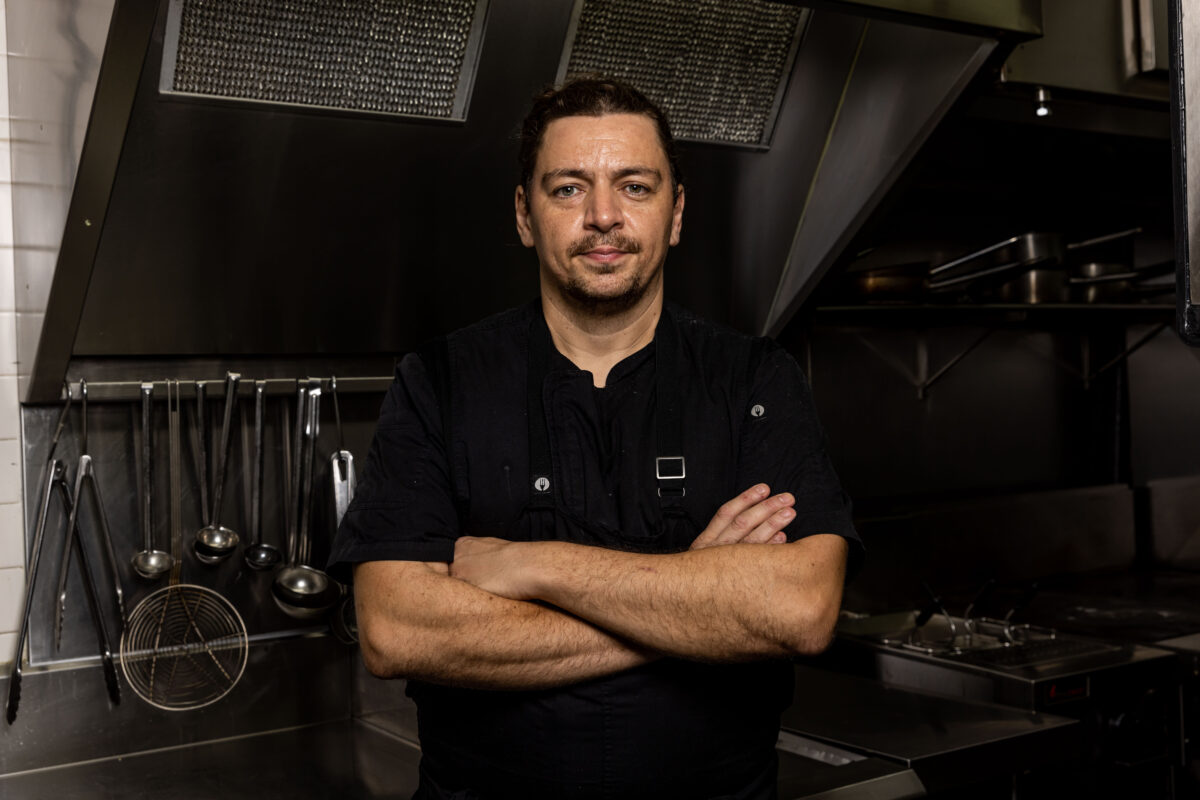 A chef with dark hair tied back, wearing a black uniform, stands with arms folded in a professional kitchen, metal utensils hanging on the wall and industrial equipment in the background.