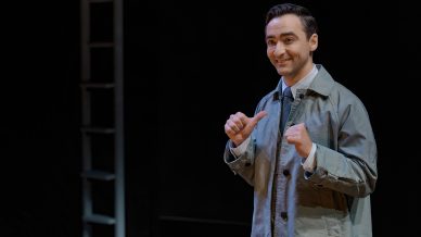 A man in a light grey coat and dress shirt stands indoors, smiling and pointing both thumbs towards himself. A metal ladder and dark background are visible behind him.