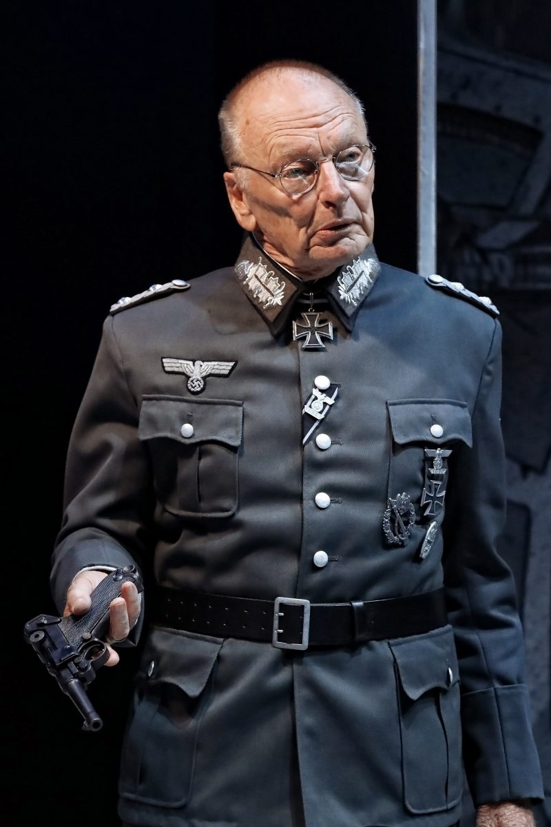 An older man in a German WWII military uniform holds a pistol in his right hand. The uniform has medals, insignia, and decorations. He wears glasses and stands against a dark background.