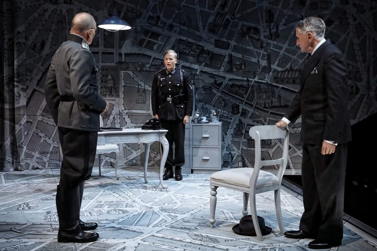 Three actors in dark uniforms and suits stand in a dramatic scene on a stage set resembling a map, with minimal furniture including a chair, desk, and filing cabinet under a hanging light.