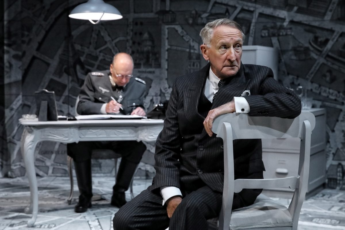 An older man in a pinstripe suit sits on a chair in the foreground, looking to the side. Behind him, a man in military uniform writes at a desk in a dimly lit, monochrome room with a map-patterned floor and walls.