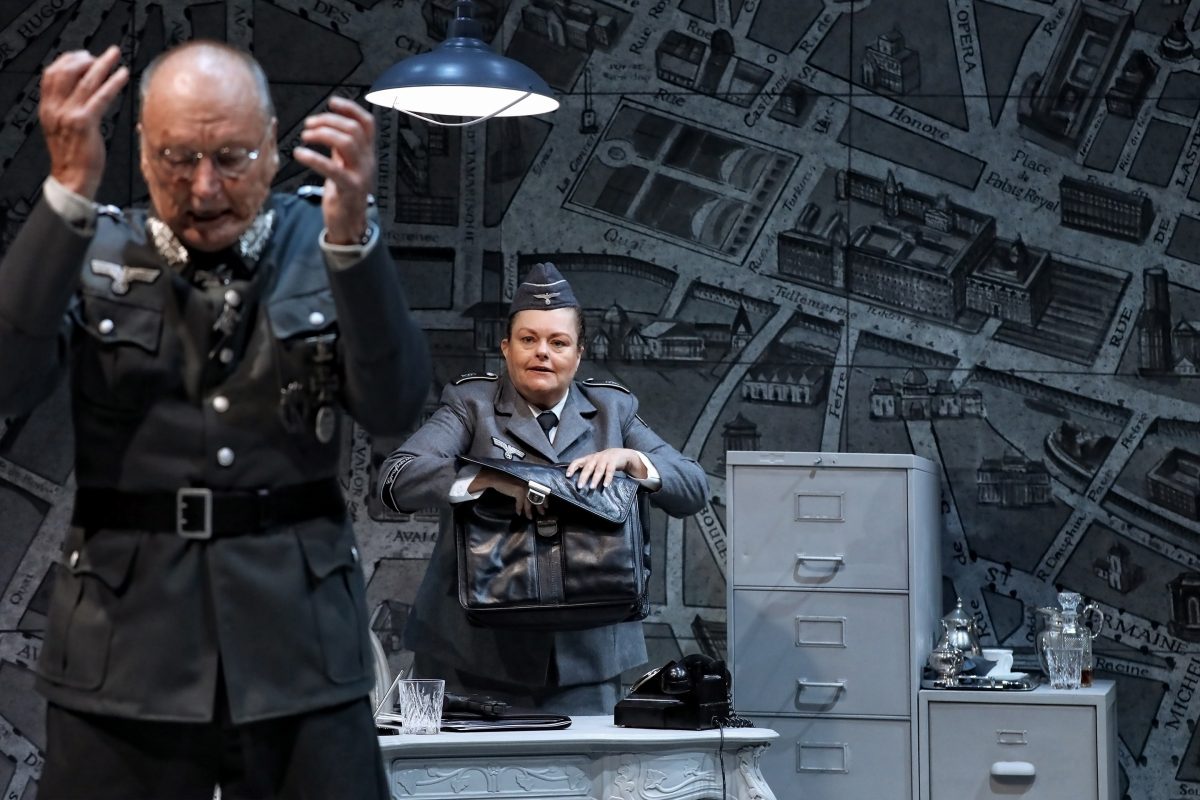 Two actors in Nazi military uniforms perform on stage; one gestures in the foreground, while the other stands behind a desk with a large bag. The set features an old city map and vintage office props.