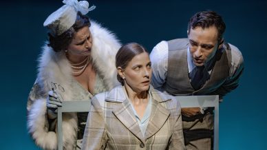 Three actors perform on stage; a woman in a fur stole and hat leans in closely, another woman in a checked suit sits looking startled, and a man in a grey suit leans in from the other side, all against a blue background.