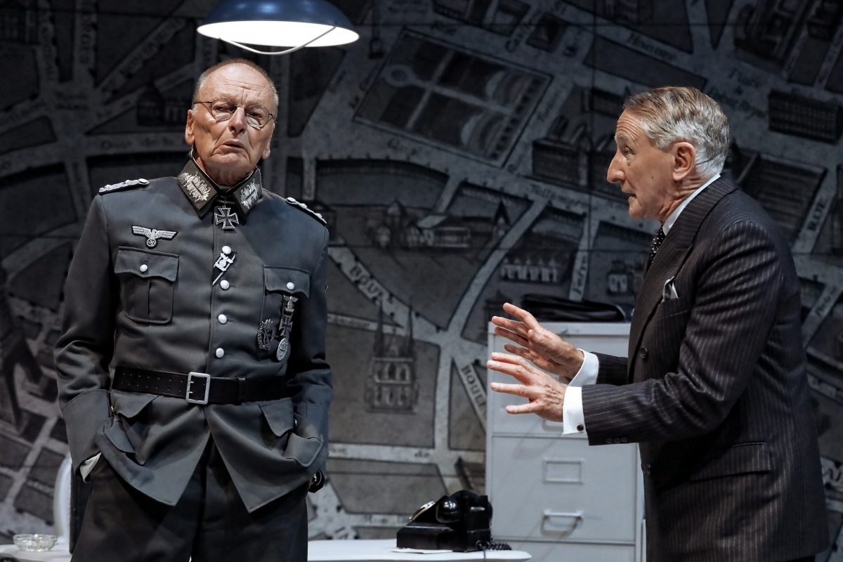 Two older men in a dramatic scene: one wears a Nazi military uniform and stands with hands in pockets, while the other, dressed in a pinstripe suit, gestures with raised hands. They stand in a dim office with a map backdrop.