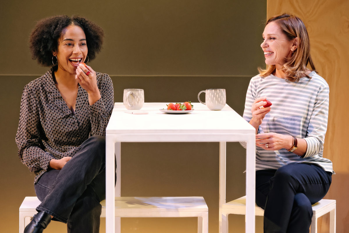 Two women sit at a white table, smiling and eating strawberries. Each has a mug, and a plate of strawberries is on the table. One woman has curly hair and wears a polka-dot shirt; the other has straight hair and a striped top.