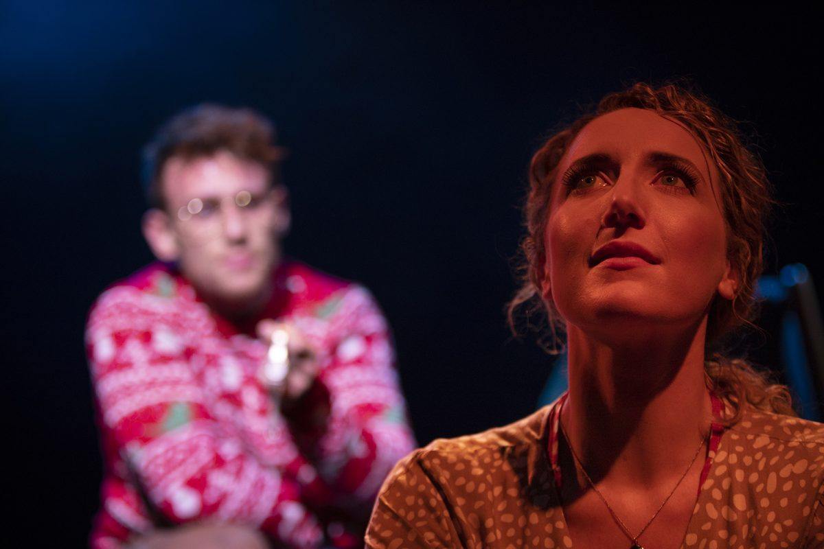 A woman in the foreground looks upwards with a serious expression, whilst a man in the background, wearing glasses and a red patterned shirt, appears out of focus and points an object forwards. The scene is dramatically lit.